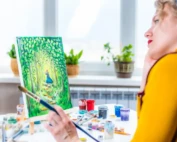 A lady is painting on a canvas while standing back and admiring her work, she is wearing a yellow sweater