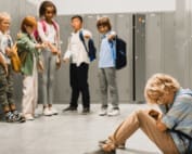 Sad crying schoolboy pupil sitting on the floor at the school hall while his classmates teenagers bully and tease him
