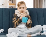 A sick, young child sitting on a couch cross-legged and drinking something to help him with his cold
