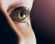 Close-up of a green eye with long eyelashes, gazing forward with soft lighting and shadow for a dramatic and introspective effect.