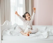 Young girl stretching and yawning in bed during a bright morning, holding a white teddy bear and surrounded by cozy white bedding.