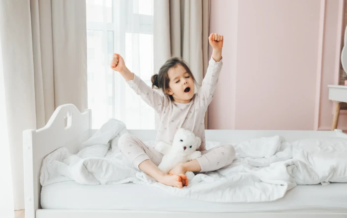 Young girl stretching and yawning in bed during a bright morning, holding a white teddy bear and surrounded by cozy white bedding.