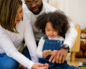 Parents sitting with their young daughter, helping her play with wooden blocks, smiling and engaged in a cozy home environment.