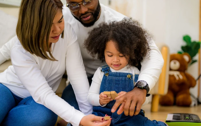 Parents sitting with their young daughter, helping her play with wooden blocks, smiling and engaged in a cozy home environment.