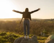 Woman standing on a rock with arms outstretched, embracing the sunset over a scenic field.