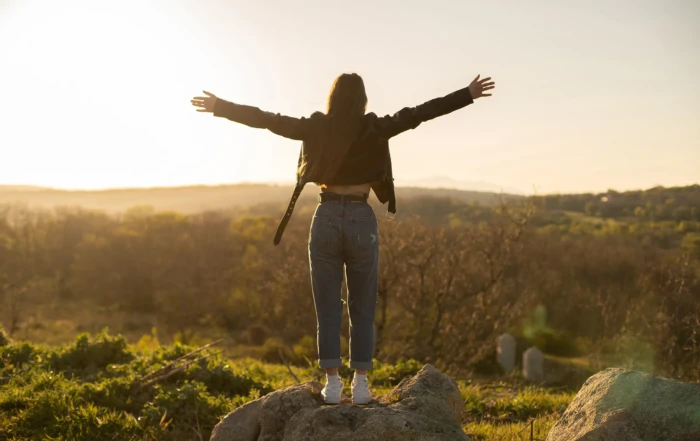 Woman standing on a rock with arms outstretched, embracing the sunset over a scenic field.