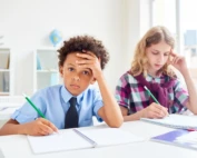Schoolboy looking frustrated while doing classwork beside a focused schoolgirl.