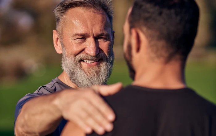 Smiling older man placing a supportive hand on a younger man's shoulder outdoors.
