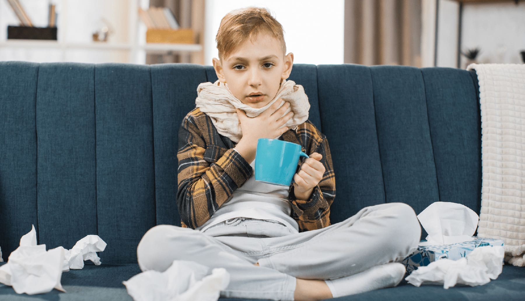 A sick, young child sitting on a couch cross-legged and drinking something to help him with his cold