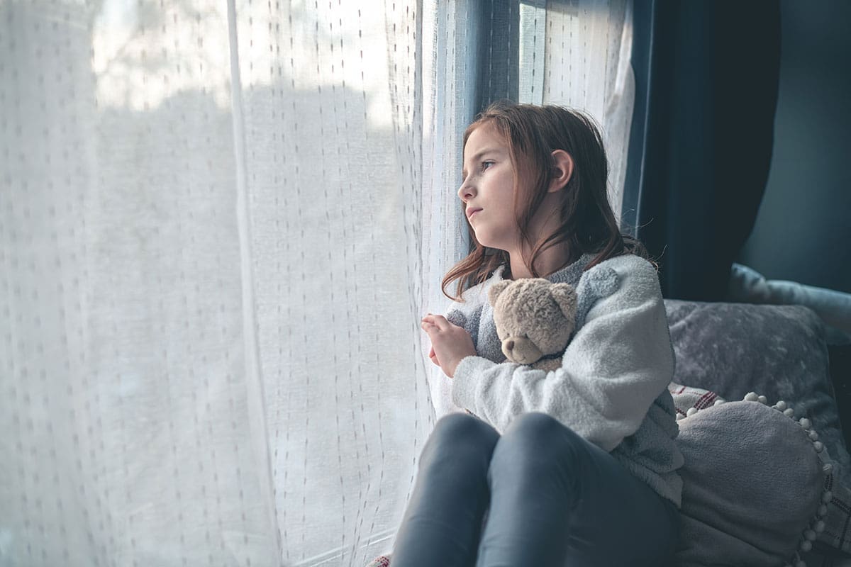 A pensive child sits and stares out the window while holding her teddy bear