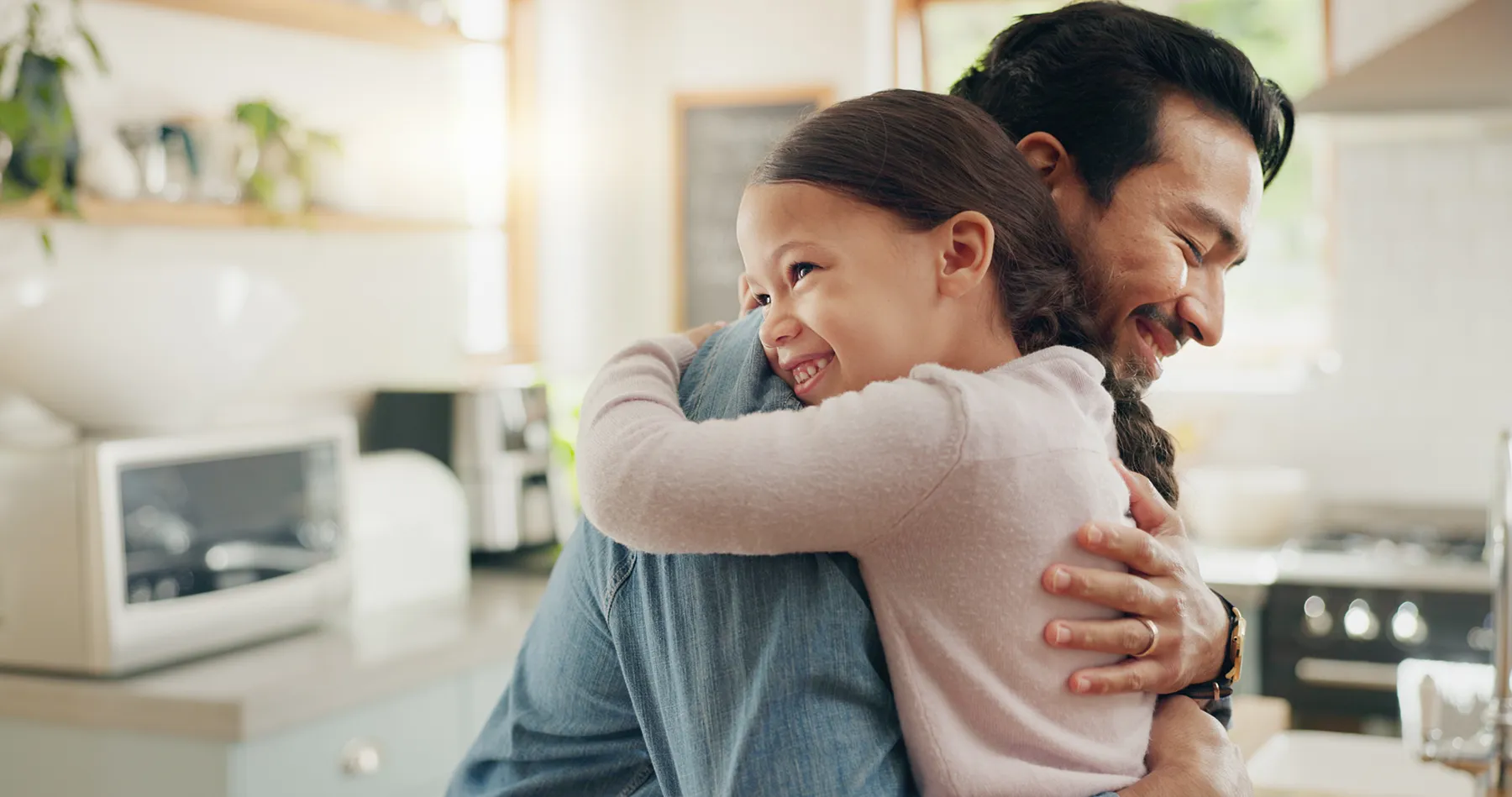 Father and daughter share a warm hug in a bright kitchen, reflecting emotional health, stress relief, and the fulfillment of a child’s emotional need for connection.
