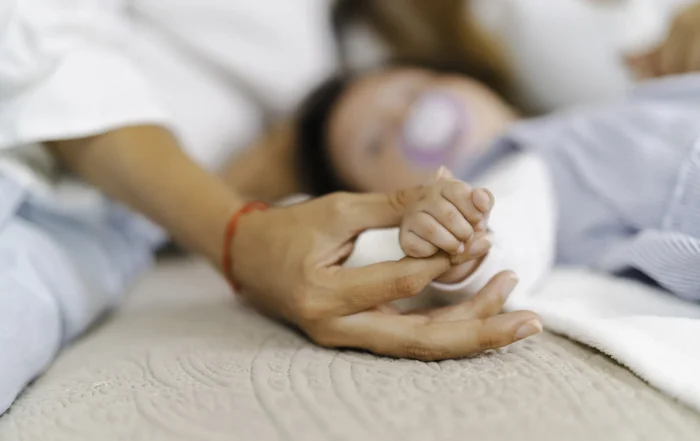 Adult gently holding a newborn baby’s hand while the baby rests on a bed with a pacifier, conveying comfort, care, and bonding.