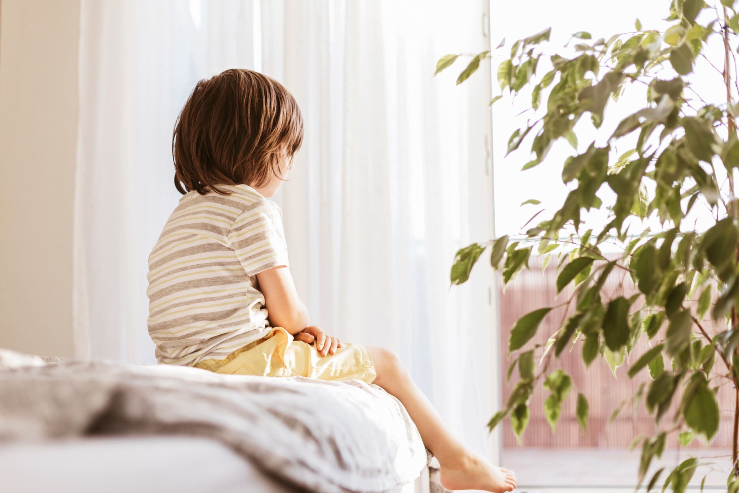 Child sitting on a bed looking out the window. Recognizing children's mental health.
