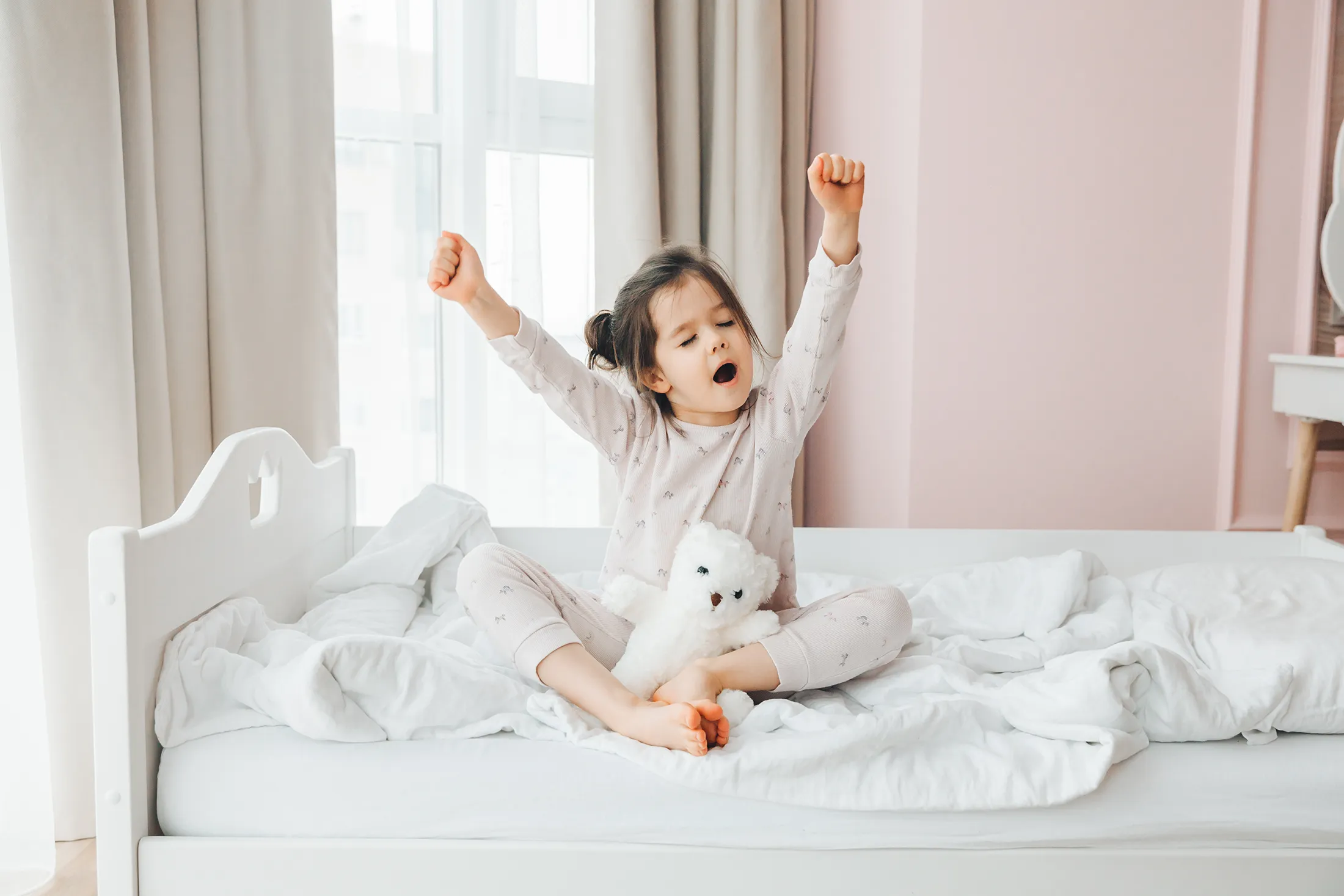 Young girl stretching and yawning in bed during a bright morning, holding a white teddy bear and surrounded by cozy white bedding.