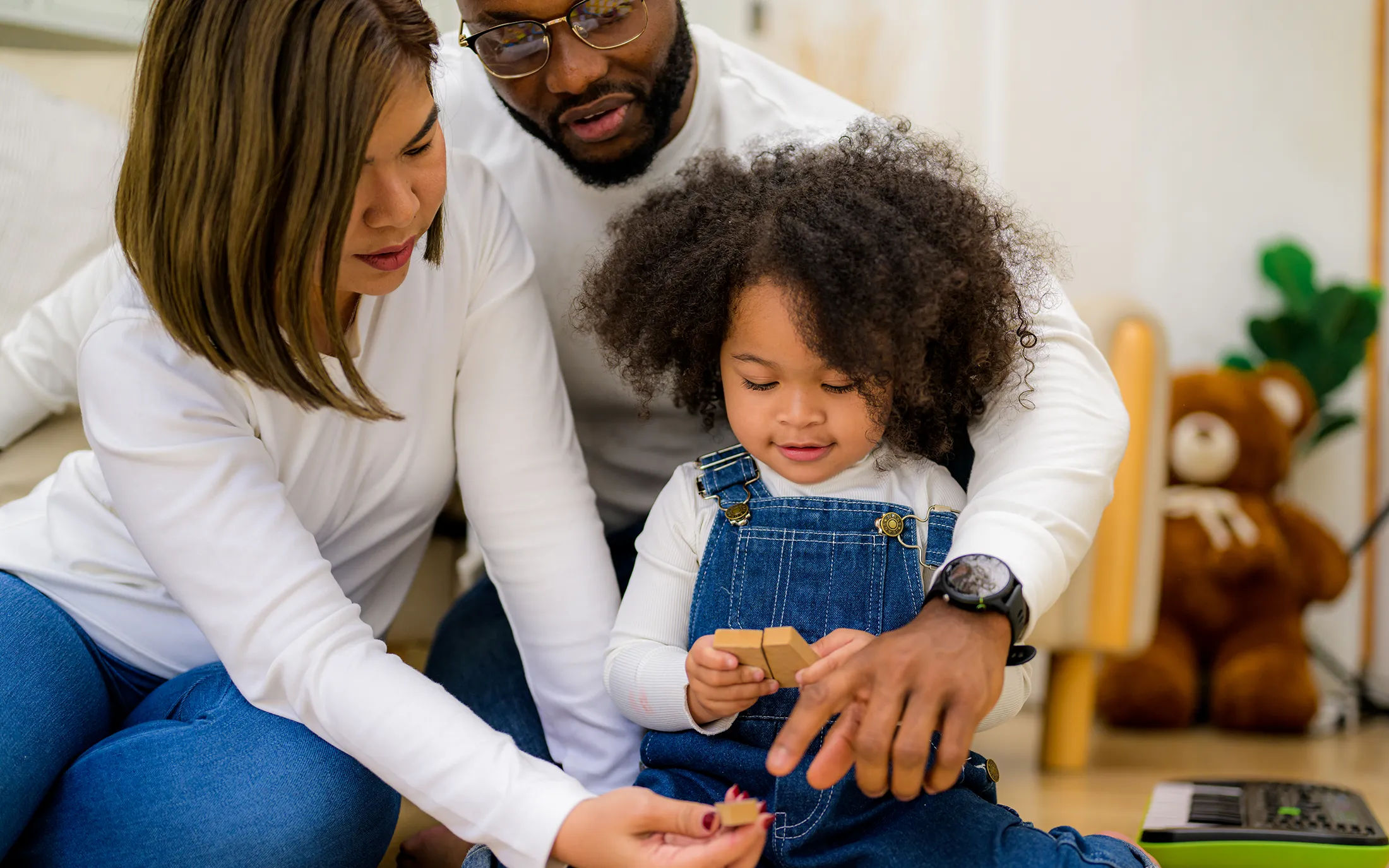 portrait-of-happy-family-father-mother-and-daughte-2024-10-11-00-00-17-utc Parents sitting with their young daughter, helping her play with wooden blocks, smiling and engaged in a cozy home environment.