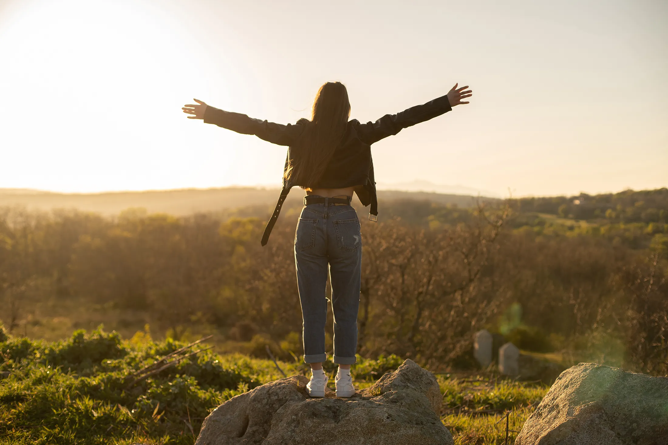 woman-with-open-arms-in-the-field-2024-12-04-11-20-28-utc Woman standing on a rock with arms outstretched, embracing the sunset over a scenic field.
