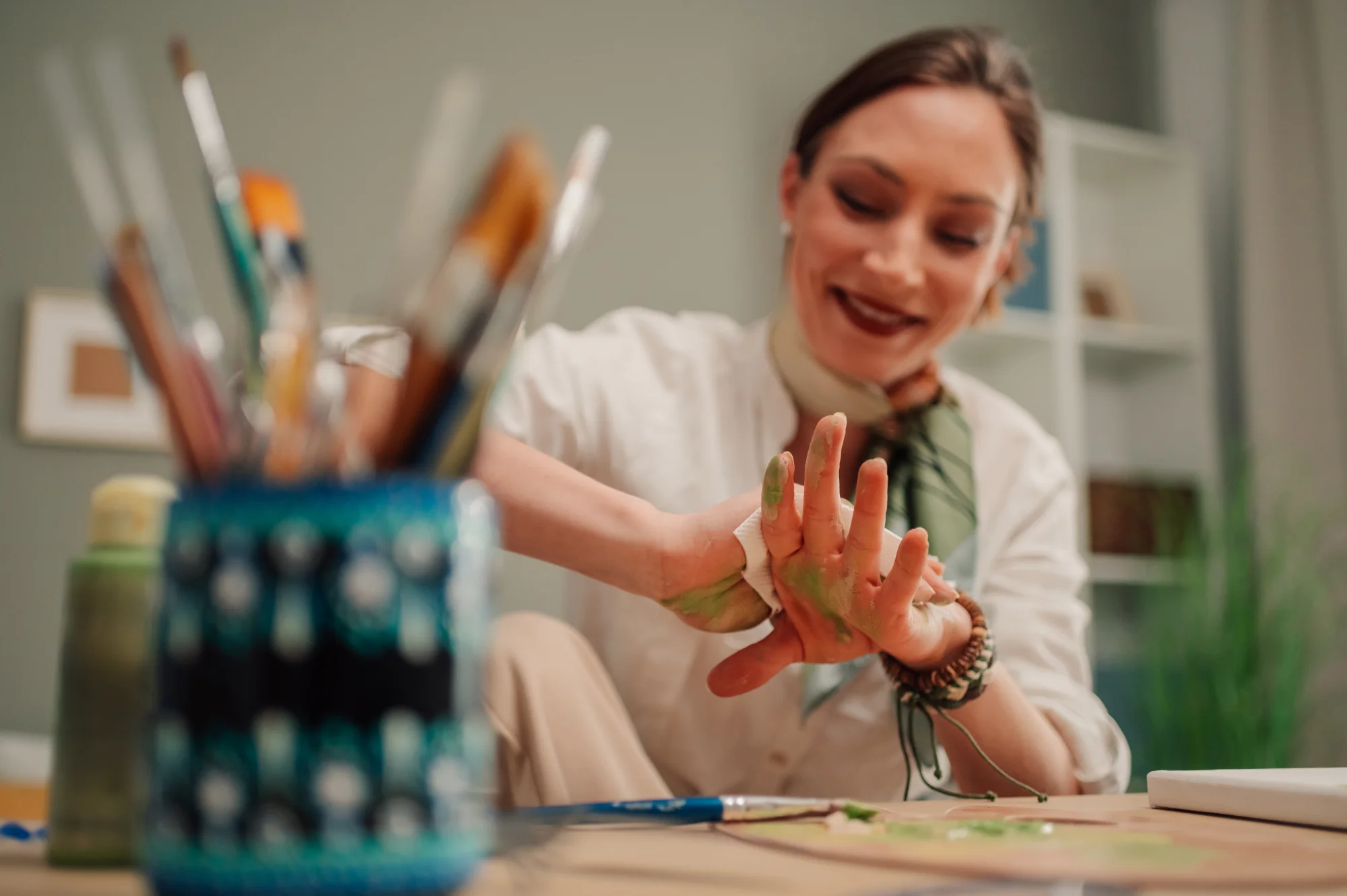 A smiling woman in a white shirt looks at her paint-covered hands during an art therapy session, with paintbrushes and art supplies visible in the foreground.