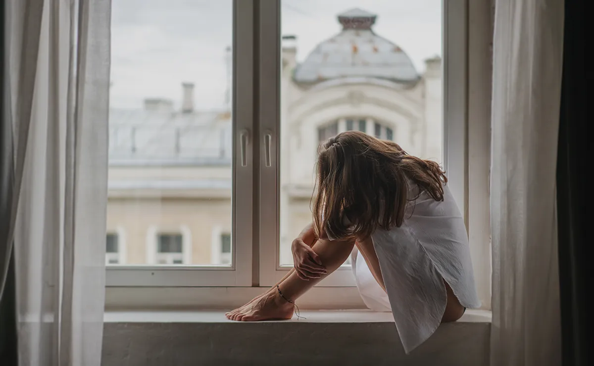 Sad woman sitting in a fetal position on a windowsill